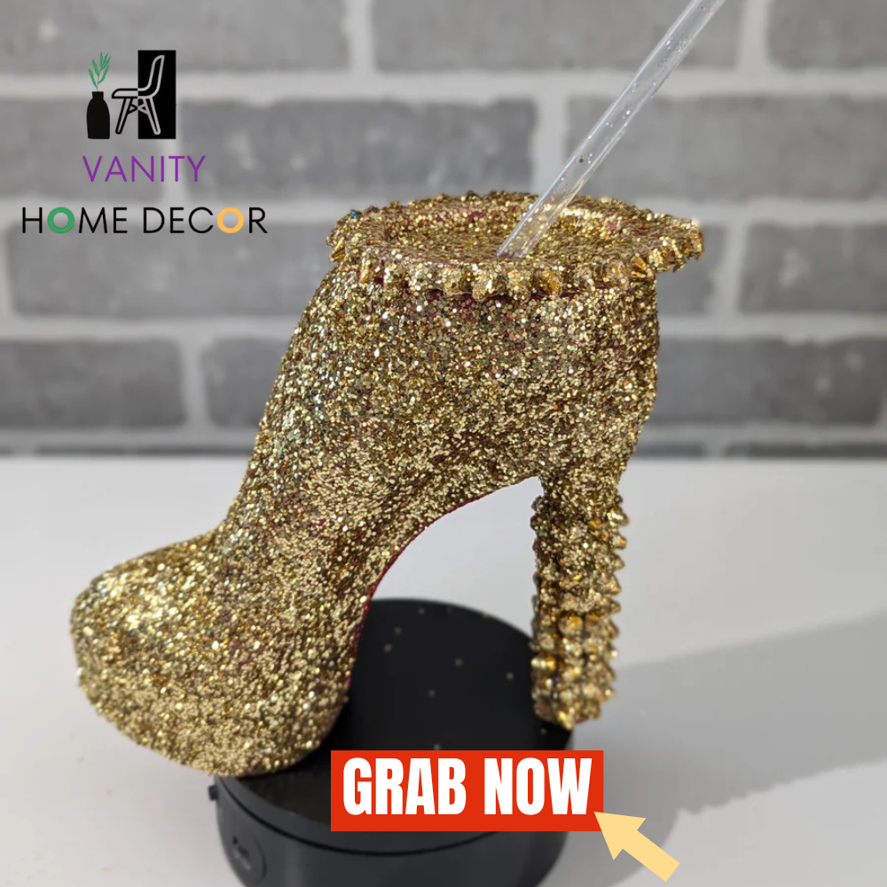 Gold glittery Spiky Stiletto Heel Tumbler Cup with a straw against a gray brick wall, branded 'Vanity Home Decor'.